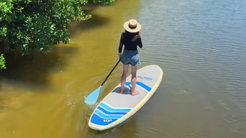 Aerial view of a woman wearing a straw hat, black top, and denim shorts paddleboarding down a narrow, calm waterway flanked by lush green mangroves.